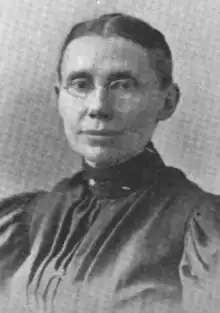 A white woman with dark hair parted center and dressed severely back to the nape; she is wearing glasses and a dark garment with a high collar, pleated bodice, and puffed sleeves