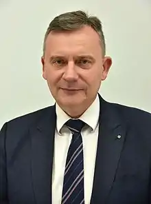 Head-and-shoulder photo of man in navy suit and a tiny green Esperanto lapel pin.