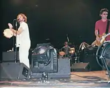 Pearl Jam on stage. Eddie Vedder sings while playing a tambourine next to a spotlight, Mike McCready plays a guitar, and Matt Cameron on the drums is seen in the background.