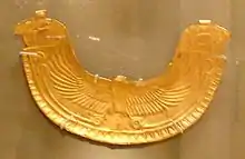 Image of a gold pectoral in the shape of an eagle spreadingh its wings. The Wings end in two Horus heads at the tip of each of the wings