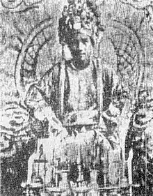 black and white photograph of a young cleanshaven man wearing an elaborate headdress and sitting on a throne. He is wearing robes and in front of him are some accoutrements on a table.