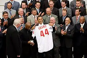 Several rows of men standing and clapping; in the front is a smiling, brown-skinned man holding a white baseball jersey with "Obama" and a large "44" in red on the rear toward camera