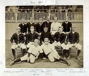 Two rows of men, one standing behind one seated, of men wearing old-style white baseball uniforms and striped pillbox caps