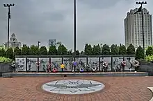 Ten large panels made of a dark stone are engraved with images from the Vietnam War. Flowers and wreaths lean against them. In front of the panels is a brick plaza with a liberty bell design.