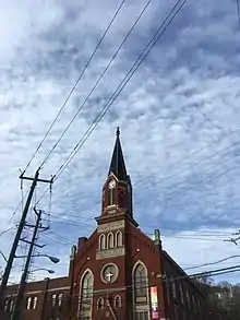 A red brick building with tan stone accents, including a stone engraved with the text: PHILIPPUS KIRCHE ERBAUT A.D. 1890. At the top of the steeple, a gilded hand points its index finger upward. The church also has a rainbow flag on its side.