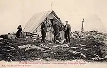 An old postcard showing several people in front of a small A-shaped wooden building