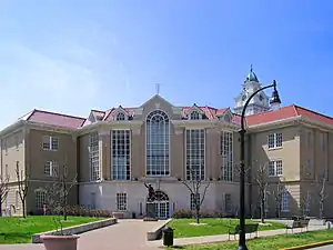 A large, brown building with towering windows, a clock tower, and a statue of a man in front