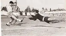 A black-and-white image taken from behind the goal net during a football match. A dark-haired player in a vertically-striped jersey and dark shorts kicks the ball with his right foot while a mustachioed goalkeeper in black dives to stop the shot. Two more players in plain, dark jerseys and white shorts are visible in the background.