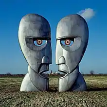 In an empty field, two metal statues resembling a human face facing each other, with a clear sky on the background.