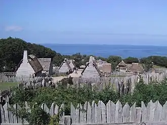 A modern-day photograph of a village consisting of small, primitive wooden houses.  Most of the houses have thatched roofs.  In the distance is a large expanse of ocean and a clear blue sky.  The village is surrounded by a wall consisting of tall, thick wooden planks.