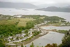 White painted cottages alongside a sea inlet, with grazing and trees behind taken from high up on a nearby crag, backed by a seascape having small islets in distance