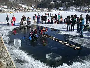  A diver is visible underwater in a hole cut in the ice cover of a small lake. Blocks of ice cut to form the hole are stacked to one side, and a second diver sits on the edge of the hole with his legs in the water. A rough wooden ladder bridges the hole. The dive site is cordoned off with a red and white tape, and other members of the support team stand to the side, with onlookers outside the cordon.