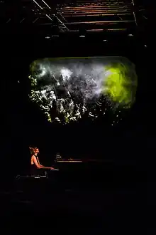 Colour photograph of Ackroyd performing in a theatre. She is seated, playing the piano, with a projection behind the piano