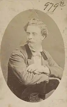 Portrait of Henri with waxed moustache, sitting backwards on a chair, 3/4 to camera, wearing a formal jacket and white bowtie.