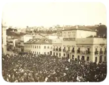 An old photograph showing a crowded square in front of a large, white, multi-storied building