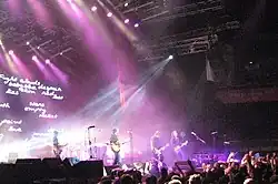 A wide shot of a stage with four musicians visible: each is playing a type of guitar. A drum kit is mid-stage but the drummer is obscured by lighting and equipment. The audience are across the front, below the stage, some have one fist raised. A photographer is centred, front of stage, another is further to the right with a camera pointing at the band, a third cameraman is left pointing into the crowd. Behind the group on the left is a large screen which depicts various words including "Fight clouds baby blue despair skin red lies lost stars ith empty rocket paint love g". Overhead lights shine down on the performers. To the right is a large sign with white writing "bring your living room to life" on red-orange background.