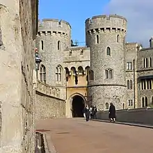 A photograph of a stone gatehouse, with two large, circular towers on each side of the gateway dominating the picture. A stone wall stretches alongside the left hand side of the picture.