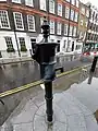 An old-fashioned water pump, painted black, with its handle removed, standing on a stone pedestal two steps above the sidewalk.