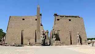 Pair of trapezoidal stone towers flanking a passage, beyond which a row of columns is visible. In front of the towers are several large statues and an obelisk.