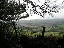 A view of hills and a patchwork of fields seen through a fence and tree.