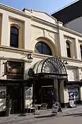 A two-storey building fronting onto a pedestrian street. The building is painted pastel-yellow and features black-framed shop fronts. An ornate black-framed semi-circular canopy protrudes above the entrance to the arcade, with gold lettering spelling out "Queen's Arcade".