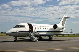 A twin-jet, high-tailplane passenger aircraft painted white above and grey below, parked on tarmac with its front stairs deployed