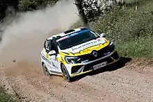 A small white-and-yellow car with a five door body is driven at speed along a gravel road.