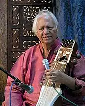 Narayan sits in front of an ornamented partition and speaks into a microphone.