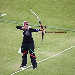 Rand Al-Mashhadani is seen getting ready to shoot in archery. She is wearing a white and pink hijab, a black long sleeve shirt, baggy black athletic pants with a green and red stripe on either leg. She holds one arm out to carry and aim the bow, and her other arm is pulled back to ready the arrow.