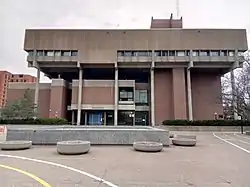 A topheavy concrete and brick building stands before a courtyard and paths.