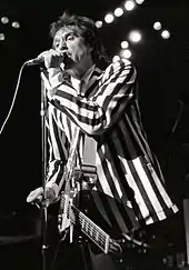 A man stands onstage with a guitar strapped across his chest. It hangs limp and unused, as he is focusing on singing into a microphone directly in front of him, which he grasps with his left hand. He wears a black-and-white, vertically striped suit.