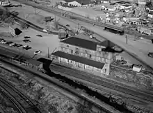 A black-and-white aerial view of a two-story railroad station. A single track, on an embankment across from the main station, has a small shelter.