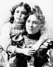 A white woman in ruffled dress, with a white girl behind her; the girl's arms are wrapped around the woman, and their faces are held close together