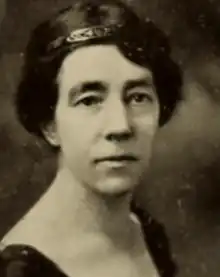 A middle-aged white woman with dark hair and brows, wearing a dark dress with a scooped neckline