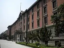 A red brick building with regular windows and a stone-faced ground floor. It is seen looking along its front; in the center there is a small entrance pavilion with a pediment at the roofline above it