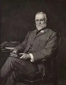 Seated photographic portrait on a chair of John Clay, holding a book and with books on a table