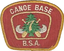 Patch with a red background and a gold border; the text Canoe Base BSA in the border; in the center a silver canoe superimposed with crossed paddles, a green pine tree and the number 7 in gold