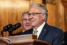 A smiling, light-skinned, bespectacled man with gray hair stands at a tan podium which has two black microphones. He wears a suit with a white-collared shirt, gold tie, and gold pin on his lapel. He stands in front of another smiling, light-skinned and gray-haired man. The second man in the background stands in front of tan, wooden walls.