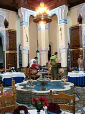 A large, open room that is filled with dining tables and chairs. In the centre of the room is a fountain shaped like a flower with eight petals. A chandelier hangs from the ceiling, which is supported by three columns at the back. A group of people eat a meal at the table closest to the columns.