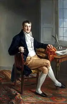 Colorful full length portrait of Meade sitting at his desk, facing the viewer. Books are strewn about.