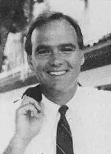 A portrait photograph featuring the suited head and shoulders of a white man, facing and smiling at the camera.