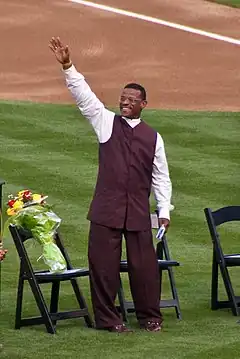 A man in brown pants, a brown vest, and white shirt raises his right hand. He is standing on grass in front of three chairs, one with a bouquet of flowers on it.