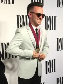 A man in a white suit and sunglasses. The text "BMI" in written several times on the wall behind him.