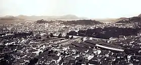 Photograph looking down over the rooftops of a large city with hills and a waterway in the far distance