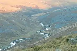 Ribbon of river in shadow reflecting the sky, bending back and forth through a valley between moorland hills of grey scree and brown and green vegetation