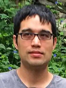 A man with short black hair and glasses seen from the neck up, where part of his gray T-shirt is visible. There are green leaves behind him