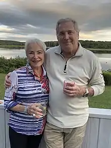 L to R: Karol L. Rose, Robert E. Pearlman are standing in front of a sunset sky and a body of water, with a green lawn visible in the near distance. They have a grey porch wall behind them and they are holding beverages.