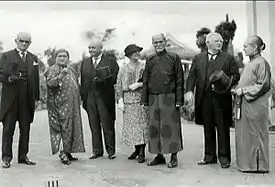  outdoor shot with four men and three women: three (Western men) wearing top hat and tails and one oriental-looking man wearing Chinese garments; two of the three women wearing Chinese robes, one wearing western dress with hat