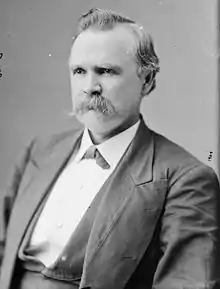 Photo shows a man with a receding hairline and a large moustache. He wears civilian clothes - a coat and white shirt.