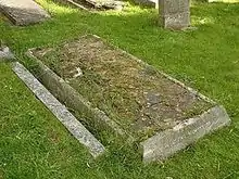 A horizontal gravestone, badly dilapidated and with grass growing among the cracks